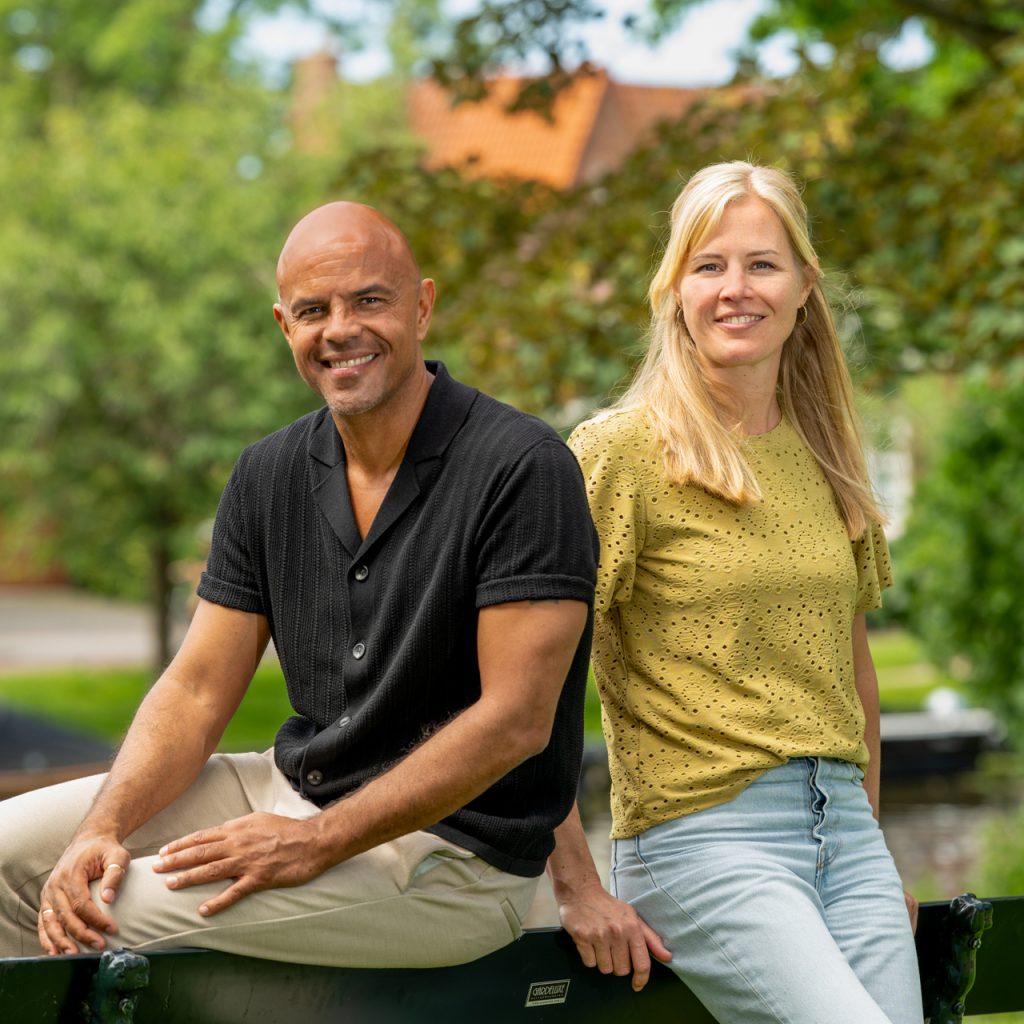 Een glimlachende man en vrouw zitten naast elkaar op een witte zitbank bij grote ramen. De man draagt een zwart overhemd en een lichte broek; de vrouw een okergele top en een lichte spijkerbroek. Gele kussens en het groene uitzicht buiten geven de ruimte een warme, lichte uitstraling.